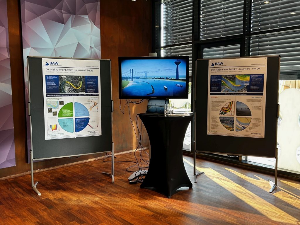 Das Bild zeigt den Stand der BAW. In der Mitte ein Bildschirm mit einem Rendering der Maßnahme in die Umgebung. Links und Rechts vom Bildschirm ist jeweils ein Poster platziert was den heutigen Zustand und einen möglichen zukünftigen Zustand zeigt und erläutert.
